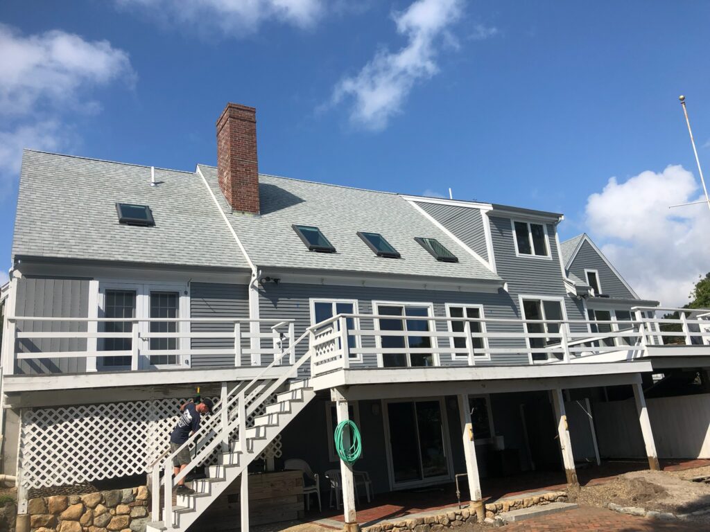 A large gray house with roofing siding, a sloped roof, and several skylights stands under a blue sky. It features a tall brick chimney, a white balcony with a staircase leading down, and a ground-level patio. A green garden hose hangs on the left side of the house.