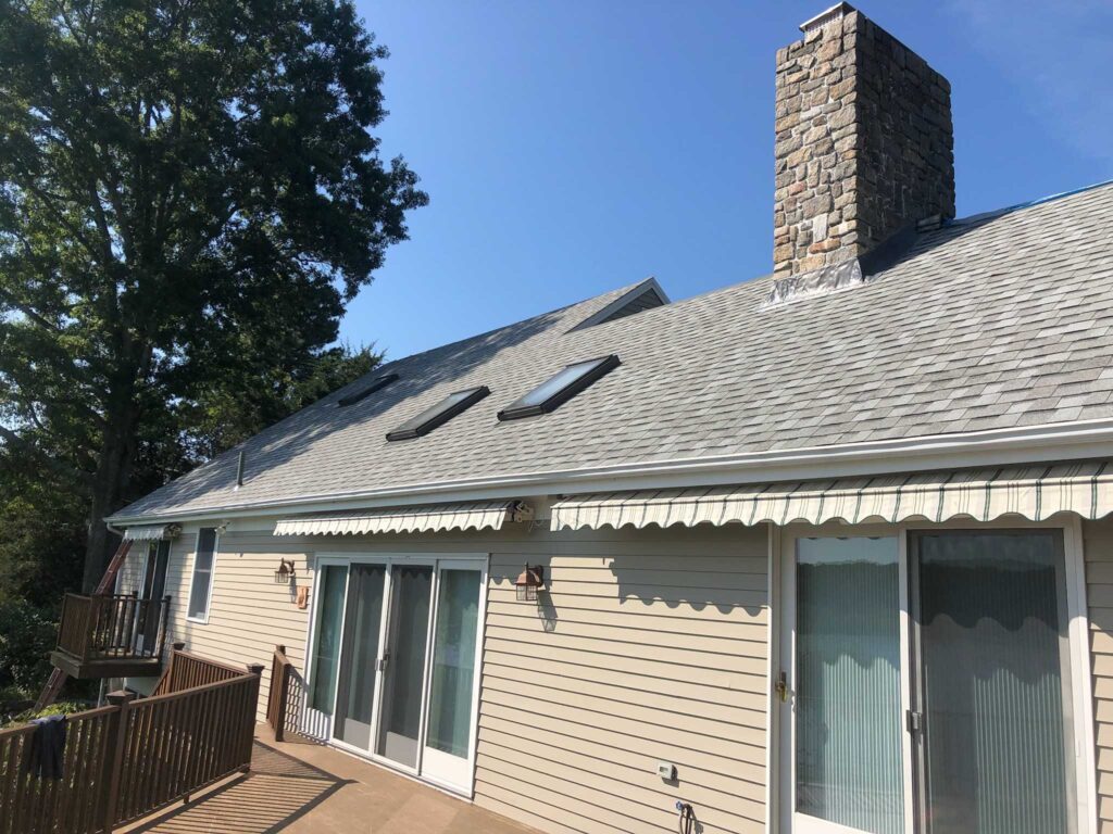 A house with beige siding, a gray shingled roof, and a large stone chimney. There are two skylights on the roof and several glass sliding doors on the side of the house. An awning extends over the doors, and a wooden deck with a railing surrounds the home. A tree is visible in the background.