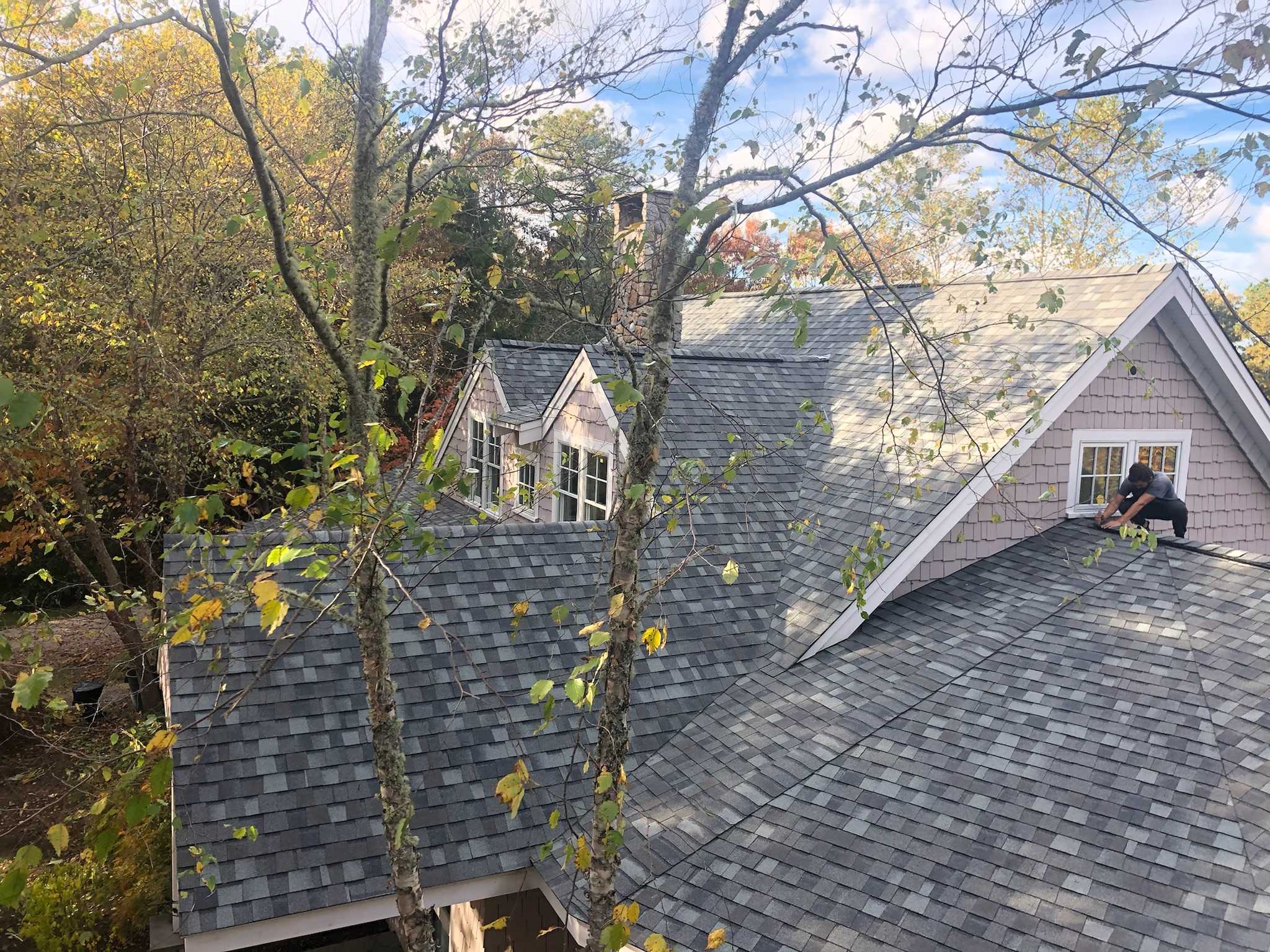 A person is working on the roof of a house surrounded by trees with autumn foliage. The shingles on the roof are gray, and the sky is partially visible through the branches. The house, complete with new roofing and siding, has multiple gables, and windows can be seen under the eaves.