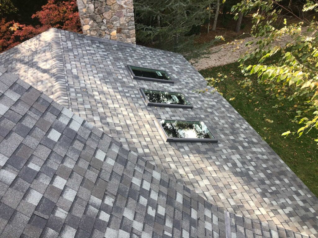 A steep, grey shingled roof features three large rectangular skylights lined up in a row. A stone chimney is visible on the left. The surrounding area has green trees and a small gravel path. The shingle pattern is a mix of light and dark grey tones.