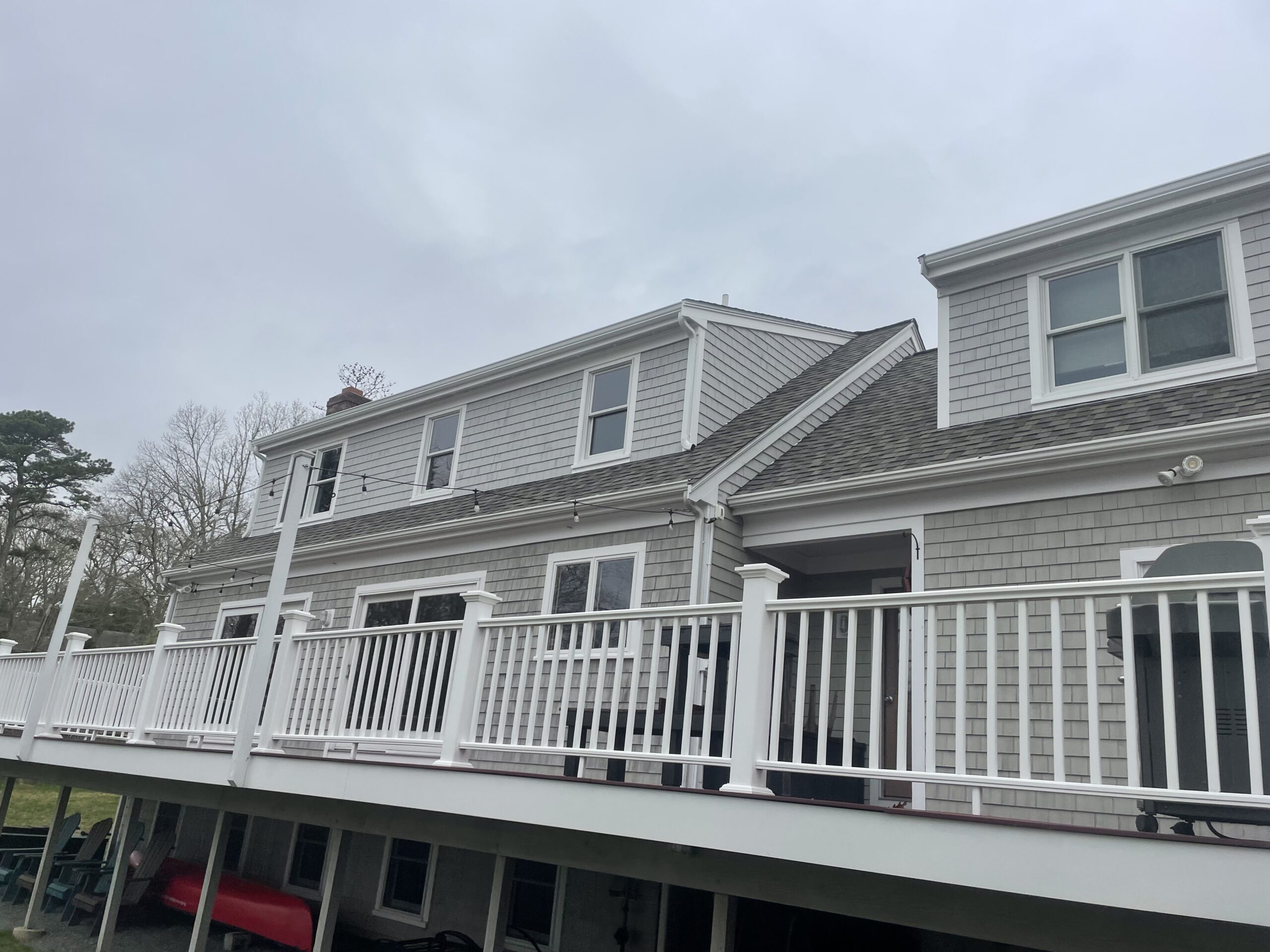 A two-story house with gray siding and white trim features a large elevated deck with white railings. The overcast sky and bare trees in the background suggest it is early spring or late fall. Outdoor furniture is visible under the deck.
