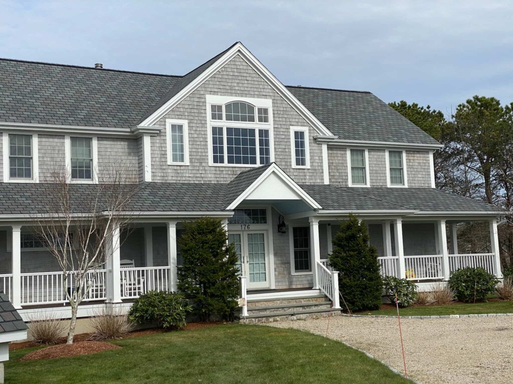A two-story gray shingle house with white trim, featuring multiple windows and a large porch with white railings. The house is surrounded by a green lawn and shrubs, with a gravel driveway leading up to the entrance. The sky is cloudy.
