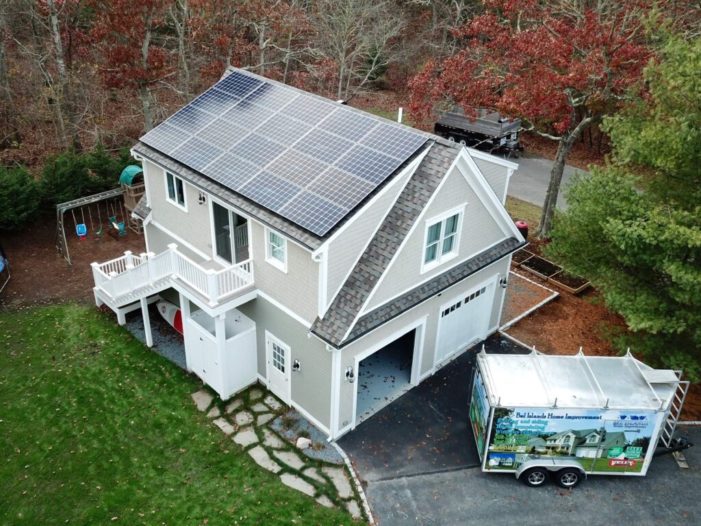 A two-story house with solar panels installed on the roof and modern roofing siding. The house, featuring a gray exterior, white trim, and a two-car garage, has a trailer with "Home Improvement" signage parked in the driveway. The surrounding yard boasts a swing set and trees adorned with autumn foliage.
