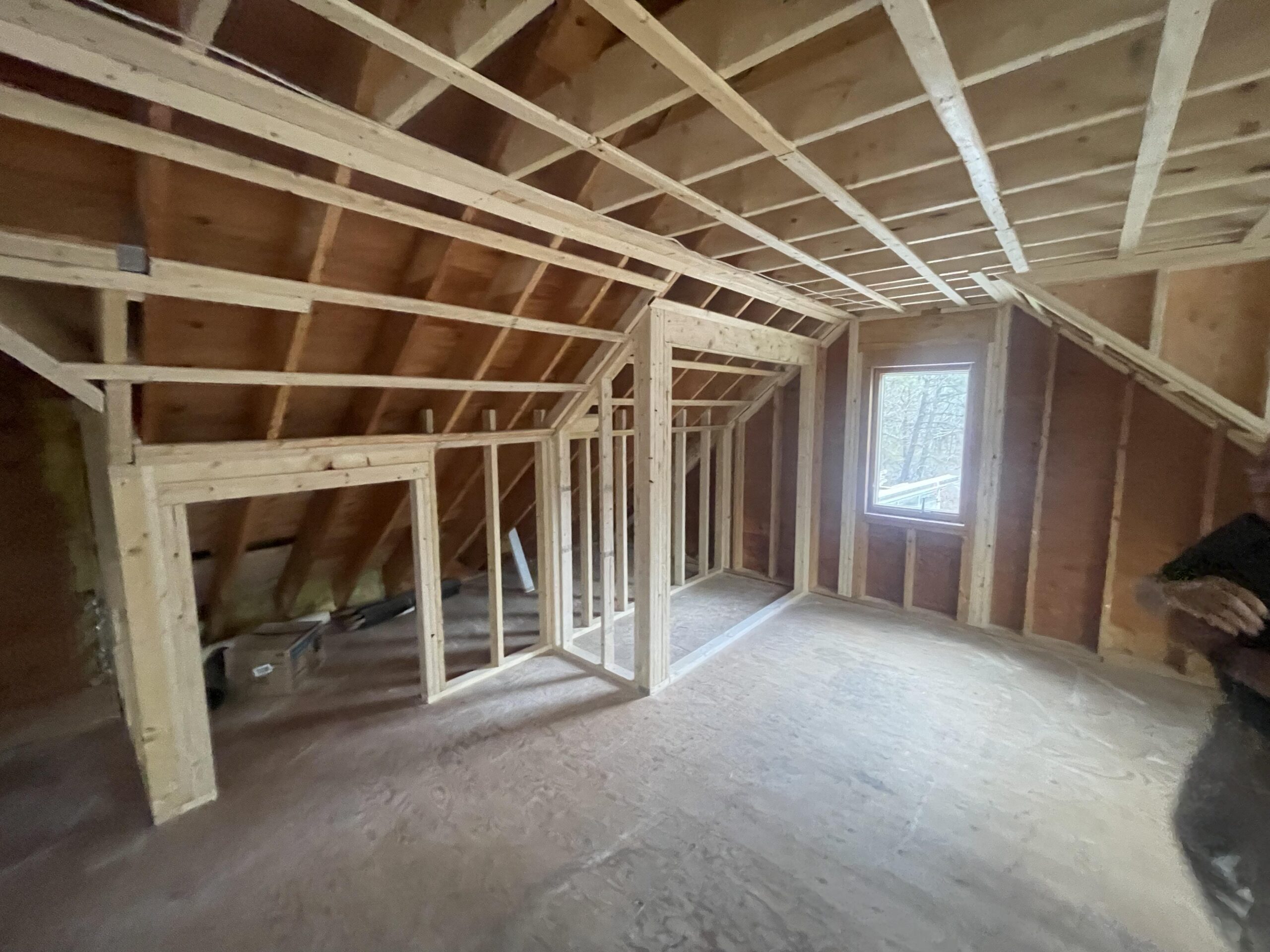 A partially constructed attic room with wooden framing, plywood flooring, and exposed beams and insulation. The room includes a window on the right side, allowing natural light to enter. Scattered construction materials and roofing siding are visible on the left.