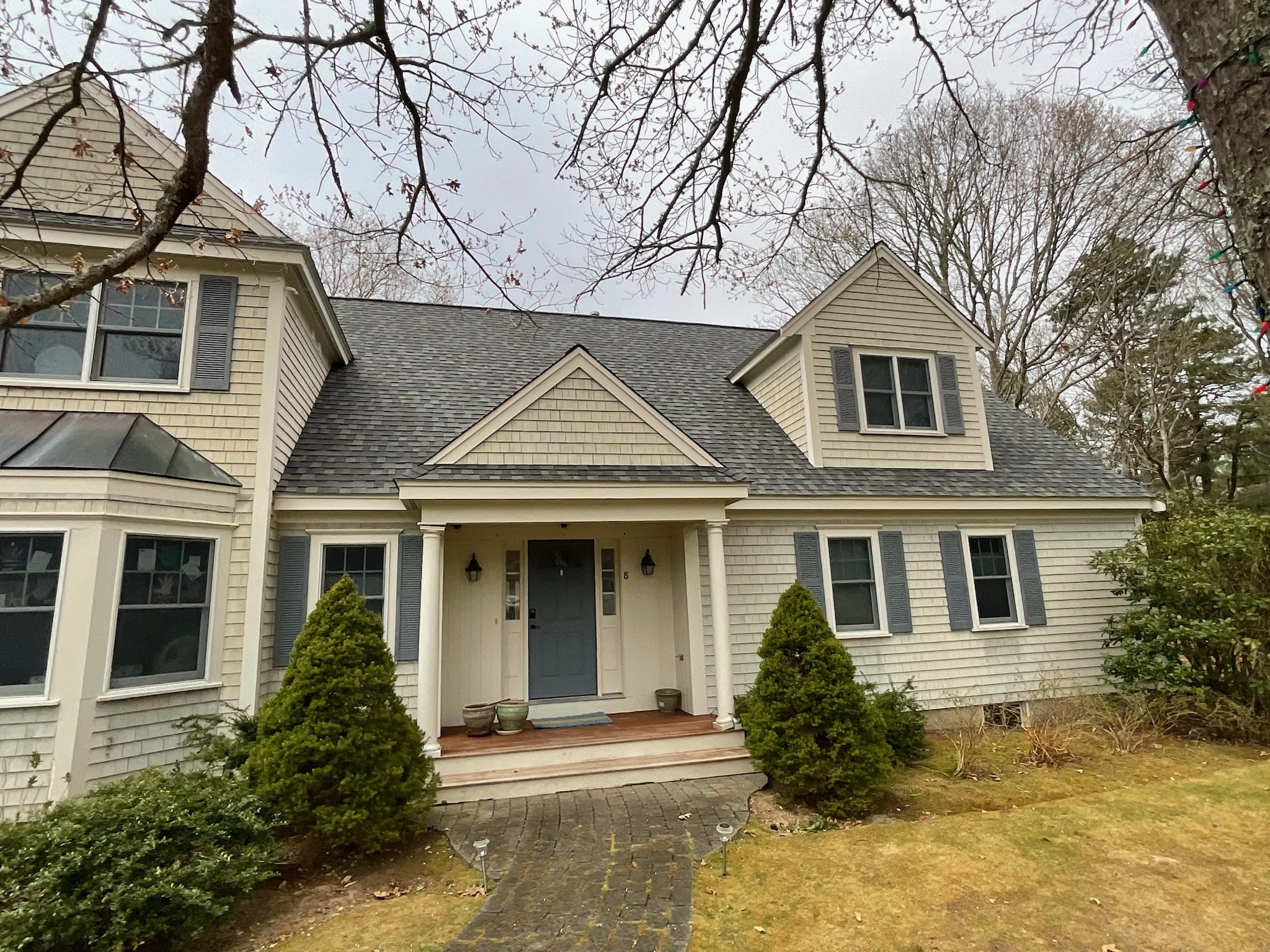 A beige two-story house with blue shutters, a dark gray shingled roof, and a welcoming blue front door. Two small trees and shrubs frame the entrance. The front yard has a stone pathway, brown grass, and bare trees in the background on a cloudy day, showcasing the quality roofing siding of the home.