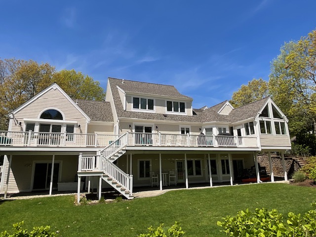 A large, multi-story house with a wide lawn and a clear blue sky overhead. The house features multiple gables, a spacious deck with railings, and numerous windows. There are stairs leading up to the deck and several trees surrounding the property.