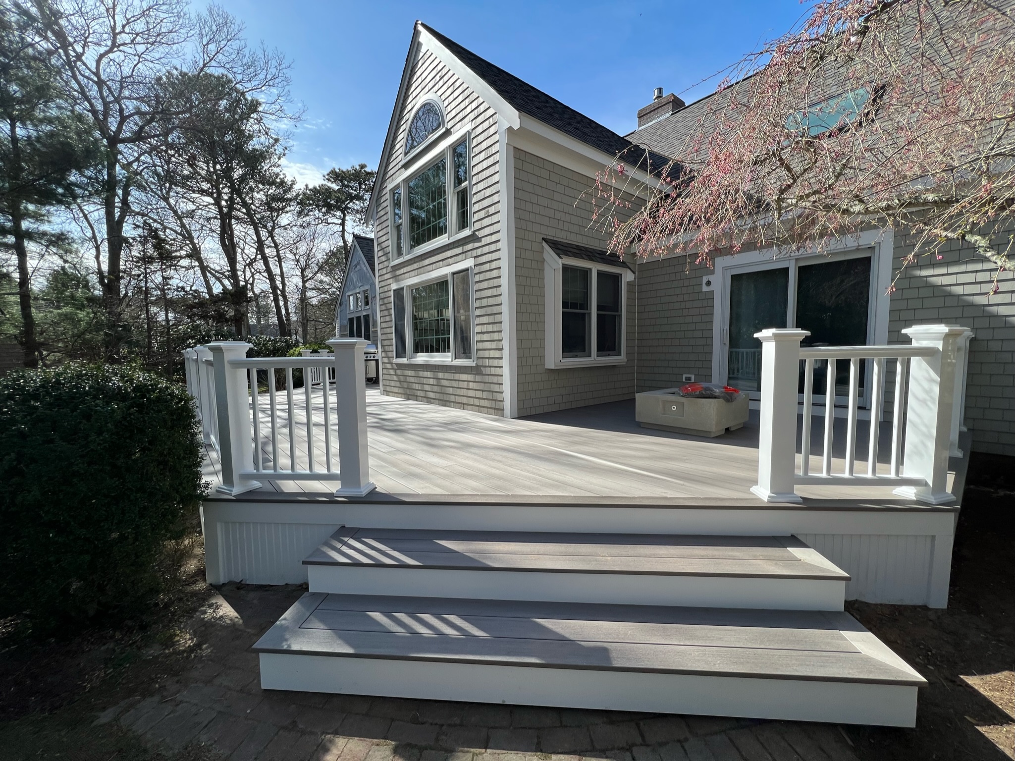A modern house with gray siding and white trim, featuring a raised deck with white railings and two steps leading down to a brick pathway. The deck, under the sturdy roofing, is furnished with a small bench and surrounded by trees and greenery on a sunny day.