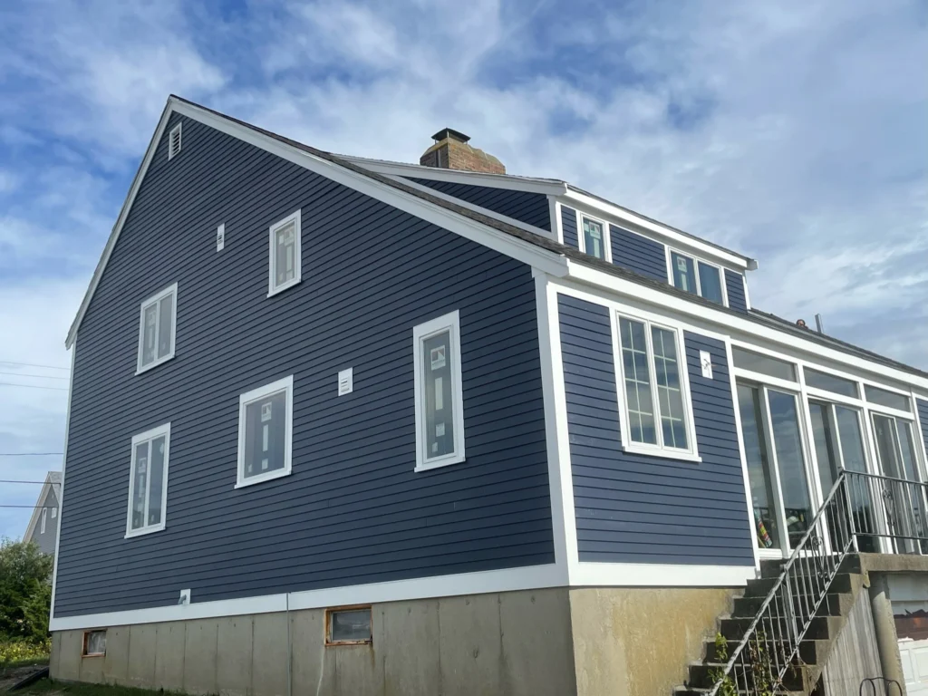 A blue two-story house with white trim is shown. The house features multiple windows, both on the main and upper floors. A staircase with a metal railing leads to the entrance on the right side. The roofing and siding complement each other beautifully against the partly cloudy sky.