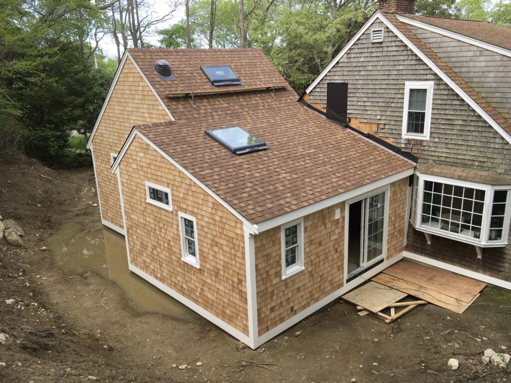 A small house extension with shingle siding and two skylights on the roof is being constructed. The attached wooden ramp leads to a sliding glass door. Muddy ground surrounds the side of the house, indicating recent excavation work. The original house with its distinct roofing is visible in the background.