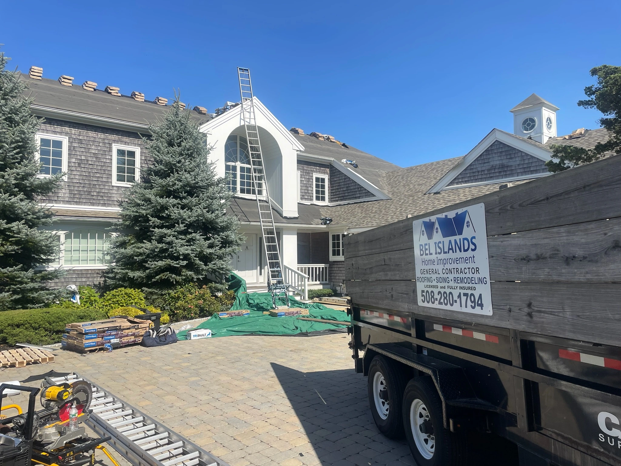 A two-story house is under renovation with roofing equipment on the roof and around the yard. A ladder leans against the house, and a sign on a trailer reads "BEL ISLANDS Home Improvement General Contractor Roofing-Siding-Remodeling" with contact details.