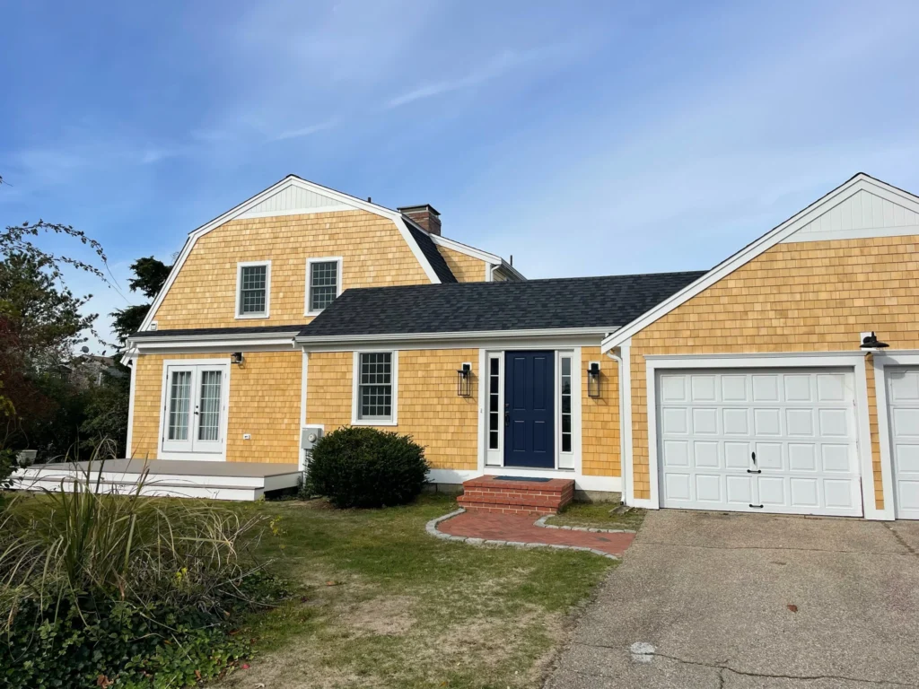 A two-story house with yellow wooden siding and white trim features a blue front door, two small windows above it, and two large windows on the ground floor. A white garage is attached to the right. The house has gray roofing and a small front garden.