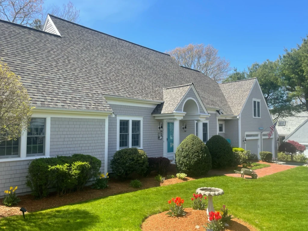 A charming single-story house with gray shingles, white trim, and blue shutters. The manicured lawn features neatly trimmed bushes, red tulips, and a birdbath. Roofing siding enhances the aesthetic near the small American flag by the side entrance. The sky is clear and blue.