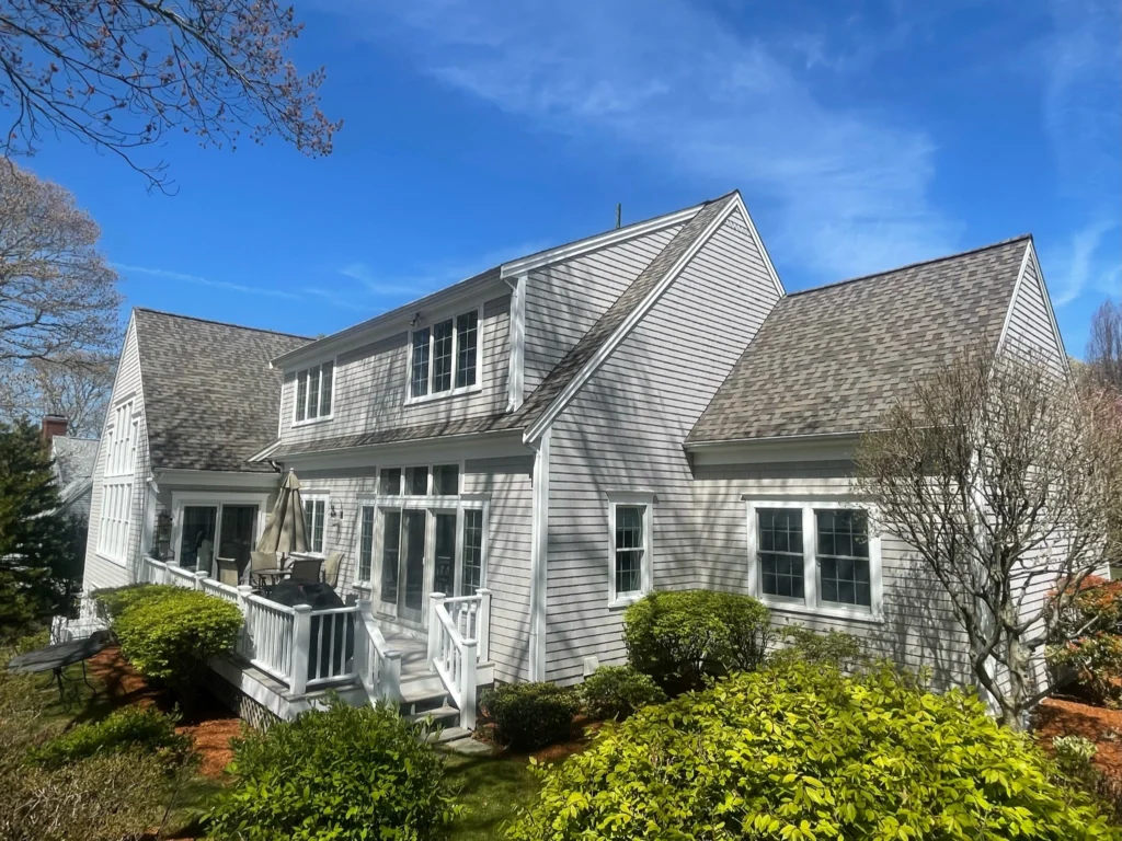 A spacious white house with gabled roofs and multiple large windows. A small patio with a table and chairs is visible, surrounded by lush green shrubs and trees. The sky is clear and bright blue, indicating a sunny day.