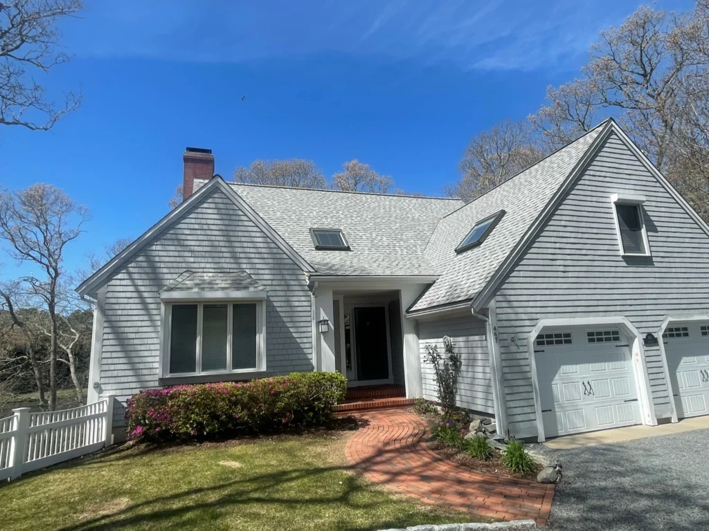 A light gray house with a steeply pitched roof and skylights, accented by white trim and new roofing siding, sits under a clear blue sky. It has a two-car garage, a front entrance with a brick pathway, green bushes, and a small white fence adorning the yard bordered by leafless trees.