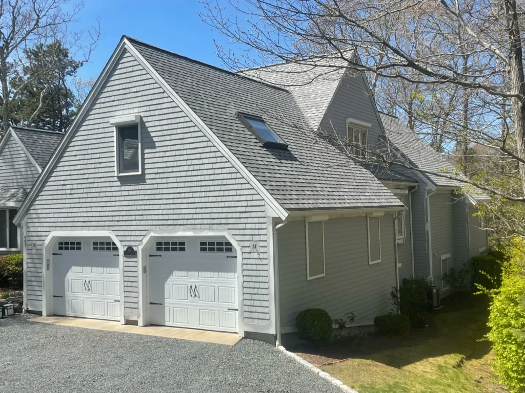 A gray, two-story house with a steeply pitched roof, two dormer windows, and a double garage featuring matching white doors. The roofing siding complements the exterior while the house is surrounded by trees, and the yard has a gravel driveway with neatly trimmed shrubbery along the sides.