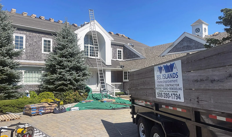 A large house is undergoing roofing work. The roof is stripped, with tiles and materials scattered on the ground. A ladder leans against the house. In the foreground, a truck with a sign reading "BEL ISLANDS Home Improvement General Contractor" is parked.
