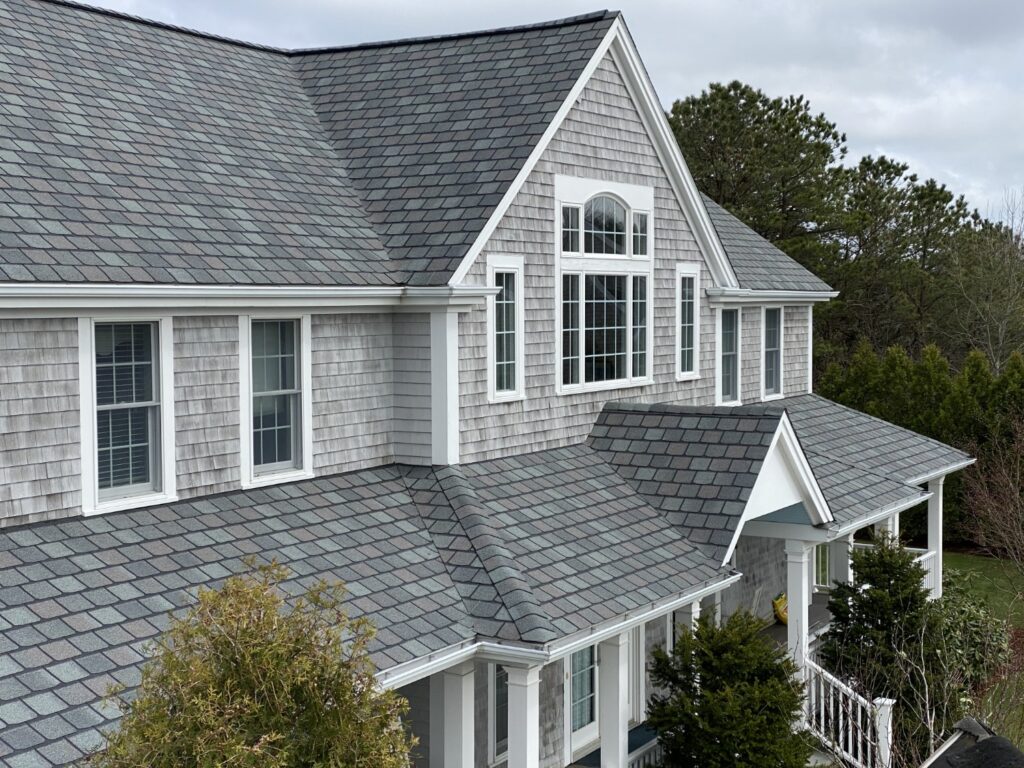 A two-story house with gray shingles and a large window on the second floor. The front porch is covered and has white railings. Surrounding the house are green shrubs and trees. The sky is cloudy.
