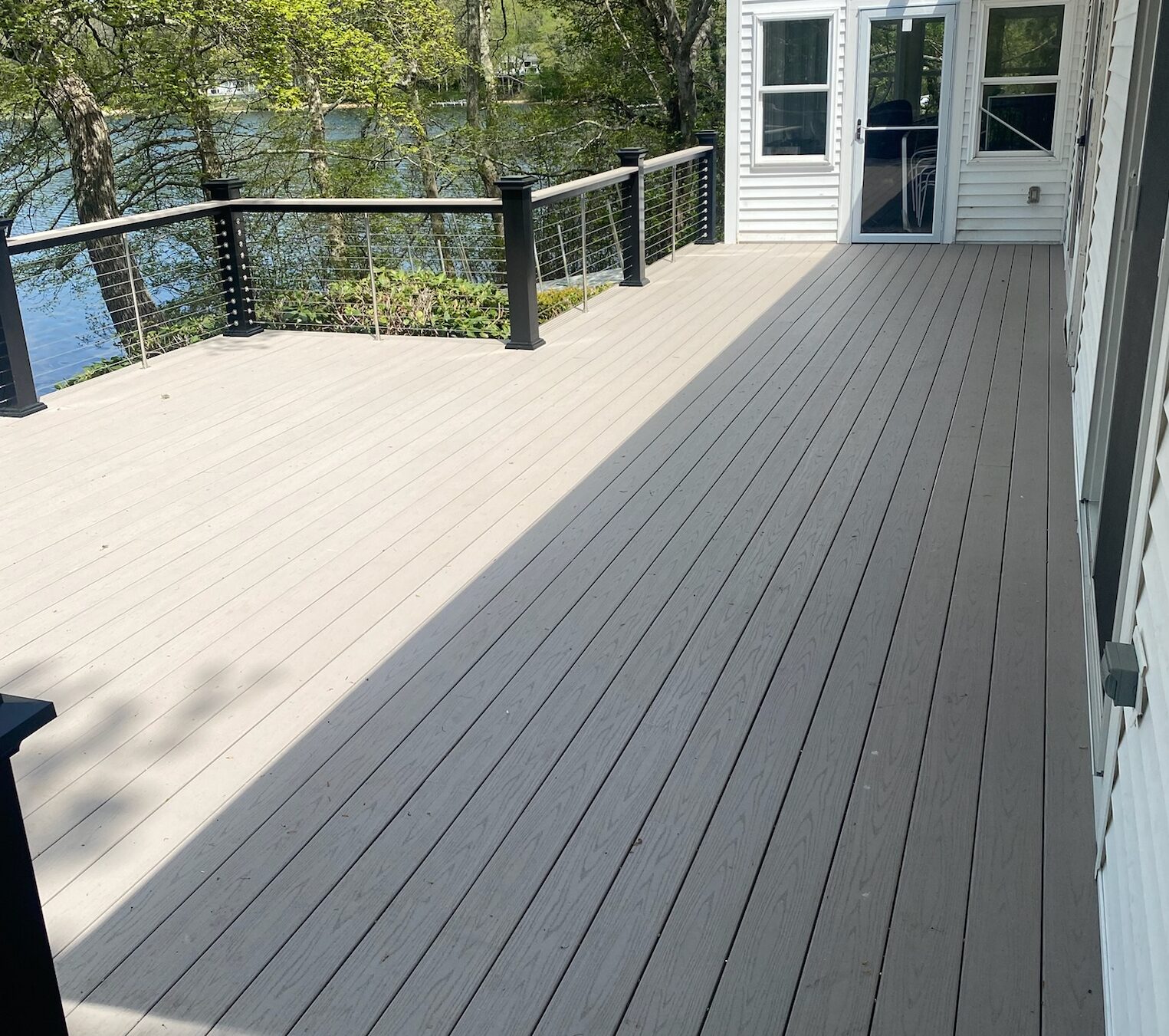 A spacious outdoor deck with light gray wooden flooring extends from a white house, bordered by a cable railing with black posts. The deck is surrounded by green trees and overlooks a scenic lake, visible in the background, under a sunny sky.