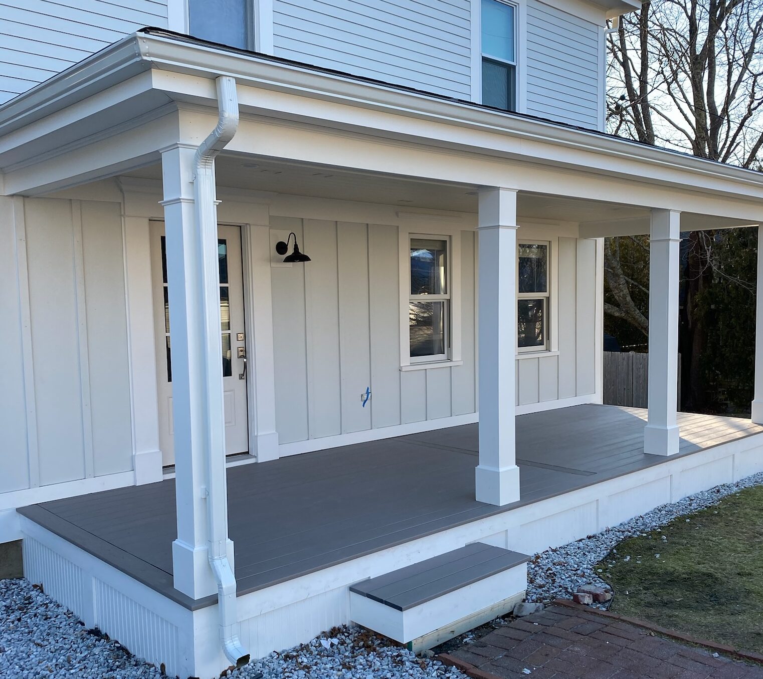 A two-story house with a white exterior and a front porch. The roofing features durable shingles, matching the white trim of the porch with its light gray wooden floor and pillars. The surrounding yard includes a brick pathway and gravel area. Multiple windows and a hanging porch light enhance its charm.
