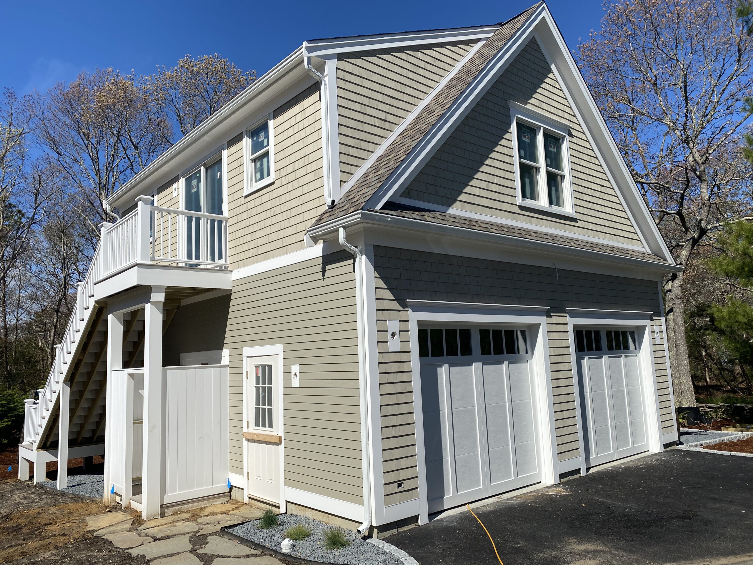 A front view of a two-story garage with light grey siding and white trim. The structure has a double garage door, an exterior staircase leading to a second-floor entrance, and small windows. The building is set against a backdrop of trees with a clear blue sky.