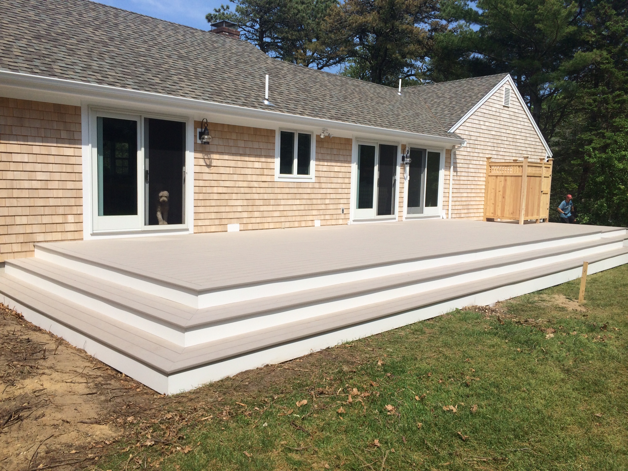 A house with light brown shingle siding has a newly built, large, light-colored wooden deck with steps. The deck spans most of the back side of the house, which features multiple sliding glass doors and a dog peeking out from one. Trees are visible in the background.
