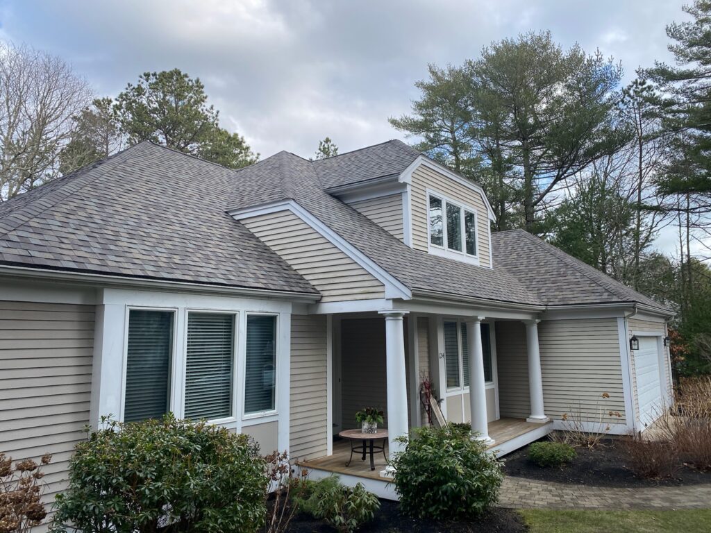 A light beige, single-story house with a steep, multi-gabled roof, white trim, and large windows. A small front porch with columns and a table with a plant at the entrance. Green shrubs and trees surround the house. The sky is partly cloudy.