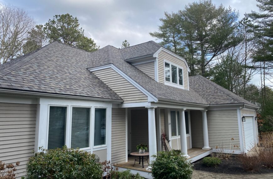 A light beige, single-story house with a steep, multi-gabled roof, white trim, and large windows. A small front porch with columns and a table with a plant at the entrance. Green shrubs and trees surround the house. The sky is partly cloudy.