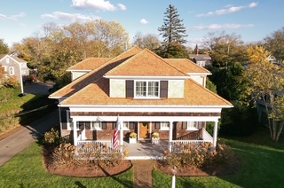 Aerial view of a two-story house with a large front porch, American flag, and manicured lawn, surrounded by trees and a sunny blue sky.