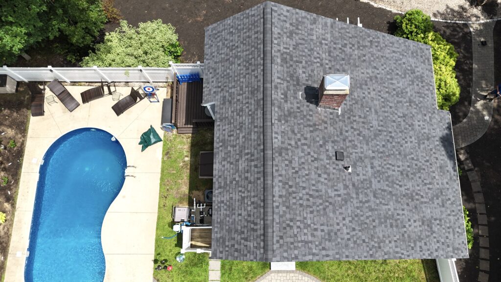 Aerial view of a house with a grey shingle roof, a fenced backyard, and a kidney-shaped swimming pool. Lawn chairs and patio furniture are near the pool, surrounded by grass and concrete patio.